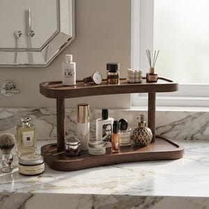 May include: A dark wood two-tier vanity tray displaying grooming products. The top shelf holds a watch, perfume, and a diffuser. The bottom shelf displays shaving cream, cologne, and skincare items. The tray is on a marble countertop.