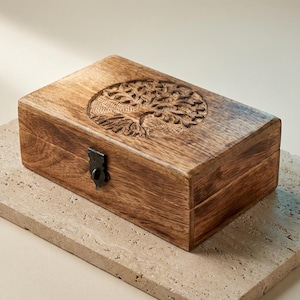Könnte beinhalten: Eine rechteckige Holzbox mit einem geschnitzten Lebensbaum-Design auf dem Deckel. Die Box ist aus hellbraunem Holz gefertigt und hat einen dunklen Metallverschluss. Die Box steht auf einer hellfarbigen Steinoberfläche.