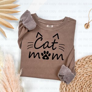 A brown sweatshirt with the text "Cat mom" printed on it, along with a cat paw print design. The sweatshirt is placed on a white background, and there are some decorative elements such as a potted plant and a woven mat in the background.