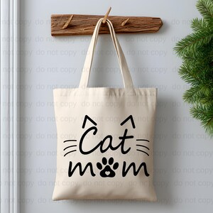 A white tote bag with the text "Cat mom" printed on it, hanging on a wooden coat rack.