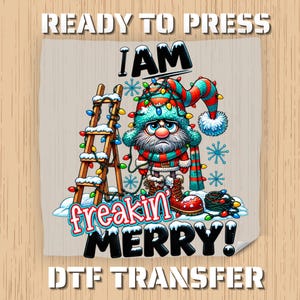 May include: A DTF transfer featuring a grumpy gnome wearing a red and blue hat and scarf, tangled in colorful Christmas lights next to a wooden ladder. The text reads READY TO PRESS, I AM freakin' MERRY!, and DTF TRANSFER in bold lettering.
