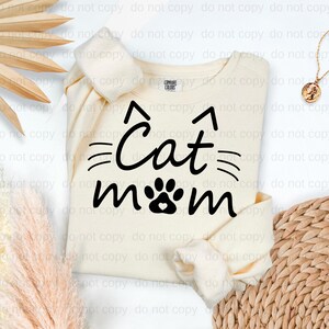 A white t-shirt with the text "Cat mom" printed on it, along with a cat paw print design. The t-shirt is placed on a white background, and there are some decorative elements such as a woven basket and a potted plant in the background.