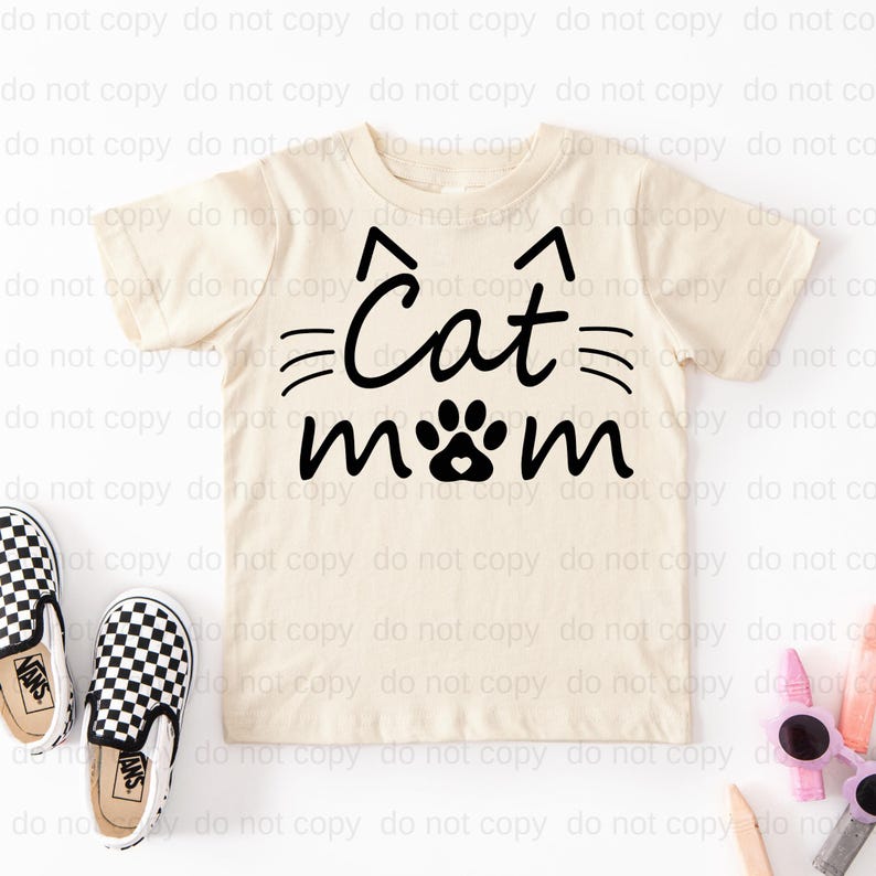 A beige t-shirt with the text "Cat mom" printed on it, along with a cat paw and paw print design. The t-shirt is placed on a white background, and there are a pair of black and white checkered shoes and some other miscellaneous items, such as sunglasses, placed next to it.