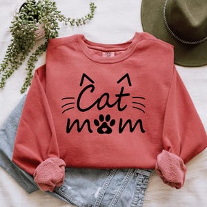 A red sweatshirt with the text "Cat mom" printed on it, along with a cat paw print, is displayed on a bed. There is also a green hat and some plants in the background.
