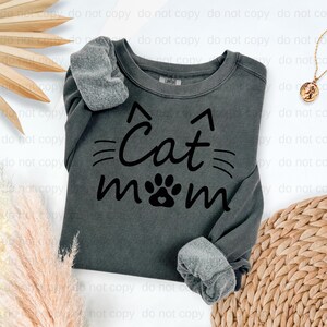 A gray sweatshirt with the text "Cat mom" printed on it, along with cat paw graphics, is displayed on a white background.
