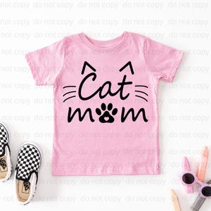 A pink t-shirt with the text "Cat mom" printed on it, along with a cat paw print design. The t-shirt is placed on a white background, and there are a pair of black and white sneakers and some pink sunglasses nearby.