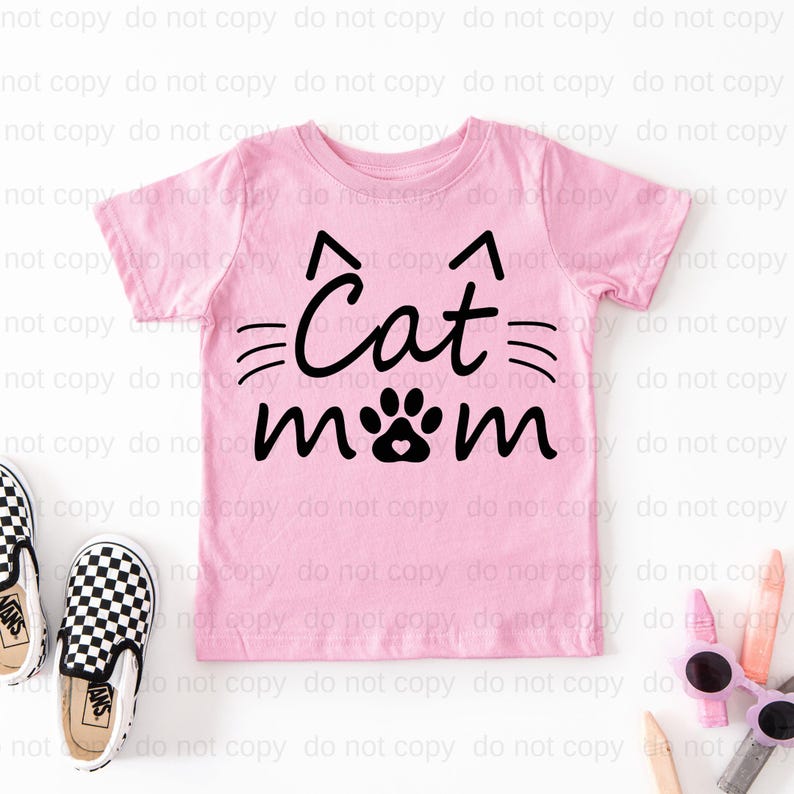 A pink t-shirt with the text "Cat mom" printed on it, along with a cat paw print design. The t-shirt is placed on a white background, and there are a pair of black and white sneakers and some pink sunglasses nearby.