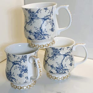 Op de afbeelding: Drie witte keramische mokken met een blauw bloemmotief en gouden rand. De mokken hebben een voetstuk en gebogen handvatten. Het bloemmotief is delicaat blauw op een crèmekleurige achtergrond. De mokken zijn op elkaar gestapeld.