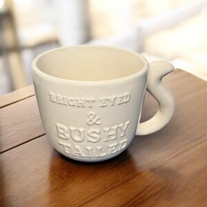 May include: Cream ceramic mug with the embossed text "BRIGHT EYED & BUSHY TAILED". The mug has a distinctive handle and is placed on a wooden surface. The mug is designed for beverages.