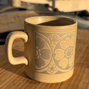 May include: A beige ceramic mug with a large C-shaped handle. The mug features a floral pattern in a lighter beige color, with a darker beige outline. The mug is sitting on a wooden surface.