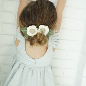 May include: A young girl with long brown hair wearing a light blue dress with a white flower hair clip. The girl is standing with her arms raised above her head.