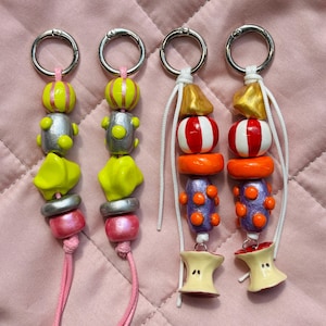 May include: Four handmade keychains with colourful beads and silver key rings. The keychains feature a variety of bead shapes and colours, including yellow, silver, pink, red, orange, purple, and gold. Each keychain has a unique design and a pink or white cord.