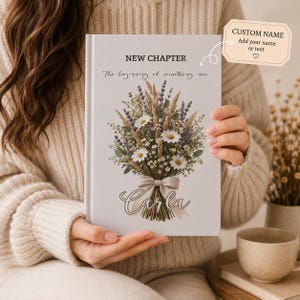 Puede incluir: Un diario de tapa dura blanco con el texto NEW CHAPTER y The beginning of something new sobre una ilustración de un ramo de flores con margaritas, lavanda y trigo atado con una cinta beige. El nombre Carla está impreso debajo del ramo.