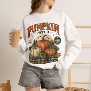 Puede incluir: Una sudadera blanca de manga larga con un estampado gráfico de tres calabazas y el texto PUMPKIN PATCH, EST. 1988, FALL VIBES ONLY y FARM FRESH LOCALLY GROWN. El diseño incluye calabazas naranjas y verdes, pinos y murciélagos.