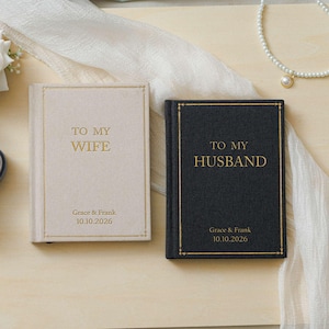 Può includere: Due libri con copertina rigida e testo in lamina d'oro. Il libro beige riporta TO MY WIFE, Grace & Frank, 10.10.2026. Il libro nero riporta TO MY HUSBAND, Grace & Frank, 10.10.2026. Un portaanelli in velluto grigio scuro contiene due anelli d'oro.
