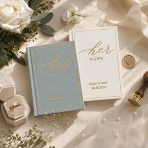 Op de afbeelding: Twee rechthoekige gelofteboeken met stoffen kaft, een lichtblauw en een wit, voorzien van goudfolie tekst met his vows en her vows, de namen Grace & Frank en de datum 10.10.2026, vergezeld van een gouden zegelstempel.
