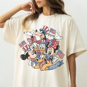 May include: Cream-colored t-shirt featuring a graphic of Mickey Mouse, Minnie Mouse, Donald Duck, Daisy Duck, Goofy, and Pluto in patriotic attire, holding American flags. The design includes the text "AMERICA 250 USA ANNIVERSARY 1776-2026".