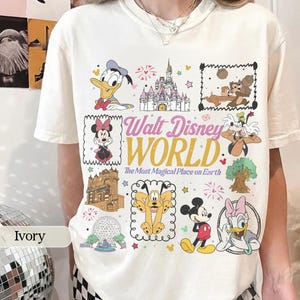 May include: Ivory-colored t-shirt featuring a colorful graphic of various Disney characters and landmarks. The design includes Donald Duck, Minnie Mouse, Chip and Dale, and the Cinderella Castle. Text reads "Walt Disney World" and "The Most Magical Place on Earth."