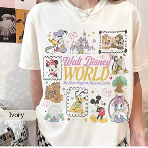 K&ouml;nnte beinhalten: Elfenbeinfarbenes T-Shirt mit einer farbenfrohen Grafik verschiedener Disney-Charaktere und Wahrzeichen, darunter Micky Maus, Minnie Maus und Cinderella's Schloss. Der Text "Walt Disney World" ist prominent dargestellt.