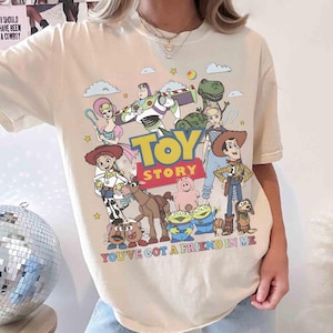 May include: Ivory-colored t-shirt featuring a colorful graphic of Toy Story characters, including Woody, Buzz Lightyear, and others. The text "TOY STORY" is prominently displayed, with "YOU'VE GOT A FRIEND IN ME" below.
