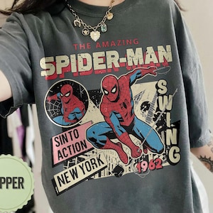 Op de afbeelding: Donkergrijs T-shirt met een vintage Spider-Man-afbeelding. Het ontwerp toont Spider-Man in actie, met de tekst "THE AMAZING SPIDER-MAN" in rood en wit. Extra tekst omvat "SIN TO ACTION" en "NEW YORK".