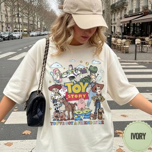 May include: Ivory-colored t-shirt featuring a colorful graphic of characters from the animated film "Toy Story." The design includes Woody, Buzz Lightyear, and other characters, with the text "TOY STORY" and "YOU'VE GOT A FRIEND IN ME."