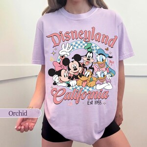 May include: Orchid-colored t-shirt featuring a Disneyland California graphic with Mickey Mouse, Minnie Mouse, Donald Duck, Goofy, and Pluto. The design includes the text "Disneyland" and "California Est 1955" in a retro style.