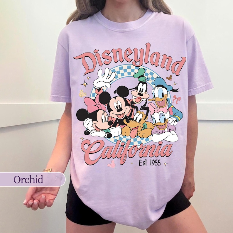 May include: Orchid-colored t-shirt featuring a Disneyland California graphic with Mickey Mouse, Minnie Mouse, Donald Duck, Goofy, and Pluto. The design includes the text "Disneyland" and "California Est 1955" in a retro style.