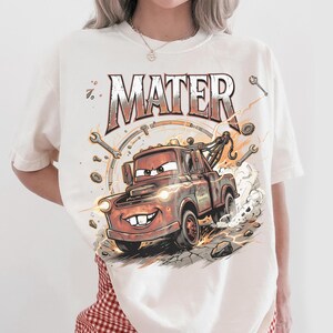 May include: Cream-colored t-shirt featuring a cartoon illustration of Mater, the tow truck from the movie Cars. The design includes the word "MATER" in a distressed font, along with wrenches and other mechanical elements.