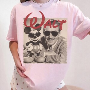 May include: Light pink t-shirt featuring a sepia-toned image of Mickey Mouse and Walt Disney wearing sunglasses. The word "Walt" is printed in red script above the image. The shirt has short sleeves and a crew neck.