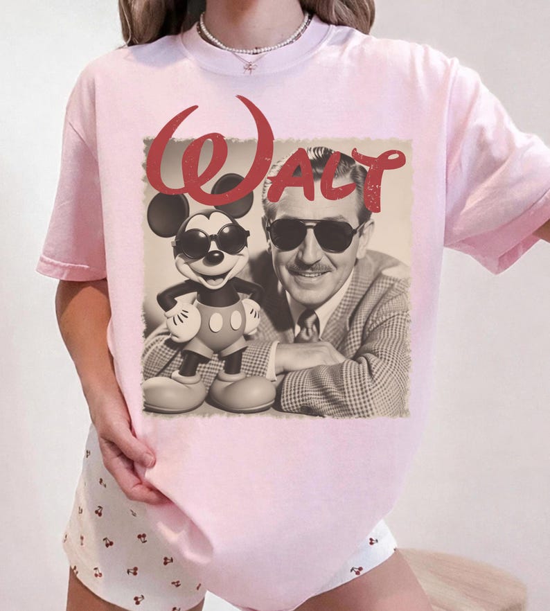 May include: Light pink t-shirt featuring a sepia-toned image of Mickey Mouse and Walt Disney wearing sunglasses. The word "Walt" is printed in red script above the image. The shirt has short sleeves and a crew neck.
