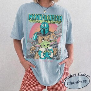 May include: A light blue t-shirt featuring a graphic print of The Mandalorian and Grogu. The design includes the text "THE MANDALORIAN" in large letters at the top and "THIS IS THE WAY" at the bottom, set against a circular pink background.