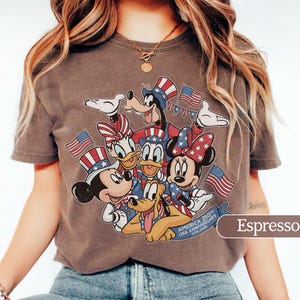 May include: Espresso-colored t-shirt featuring a graphic of Mickey Mouse, Minnie Mouse, Donald Duck, Daisy Duck, Goofy, and Pluto in patriotic attire. The design includes American flags and the text "AMERICA 250th USA ANNIVERSARY 1776-2026".