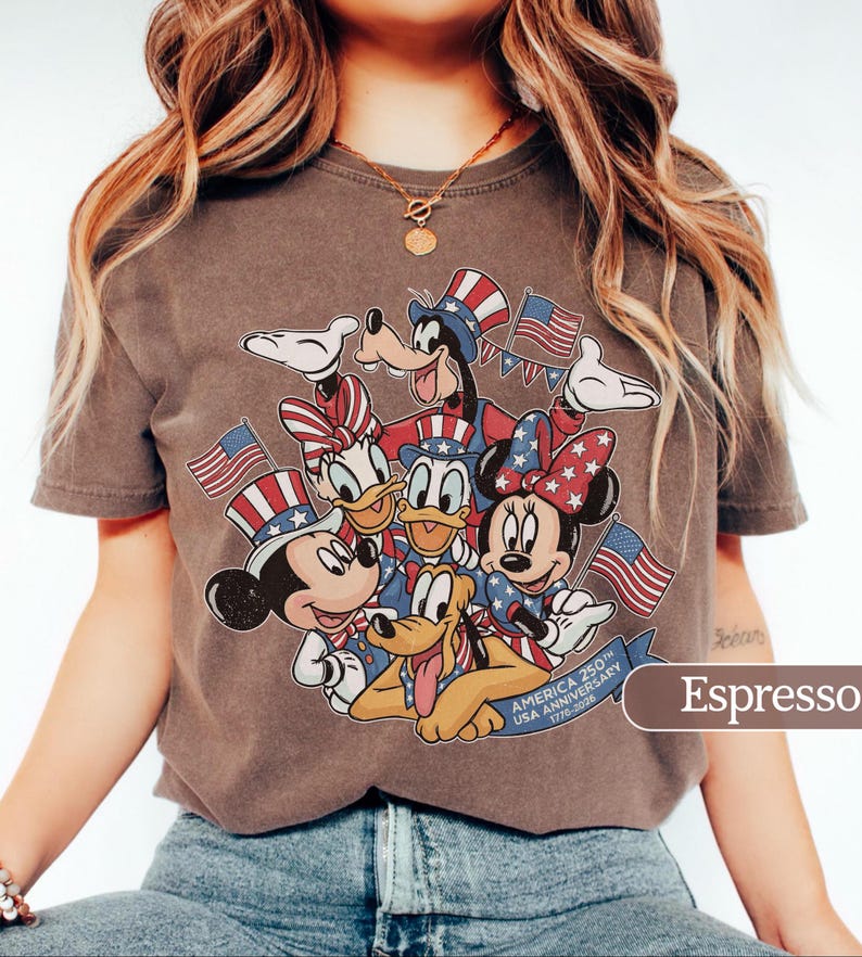 May include: Espresso-colored t-shirt featuring a graphic of Mickey Mouse, Minnie Mouse, Donald Duck, Daisy Duck, Goofy, and Pluto in patriotic attire. The design includes American flags and the text "AMERICA 250th USA ANNIVERSARY 1776-2026".
