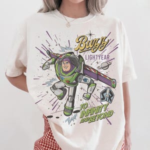 Puede incluir: Camiseta blanca con un dise&ntilde;o colorido de Buzz Lightyear de Toy Story. El dise&ntilde;o incluye el texto "Buzz Lightyear" y "To Infinity and Beyond" con un fondo tem&aacute;tico espacial.
