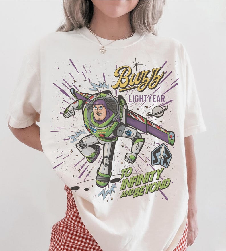 Puede incluir: Camiseta blanca con un dise&ntilde;o colorido de Buzz Lightyear de Toy Story. El dise&ntilde;o incluye el texto "Buzz Lightyear" y "To Infinity and Beyond" con un fondo tem&aacute;tico espacial.