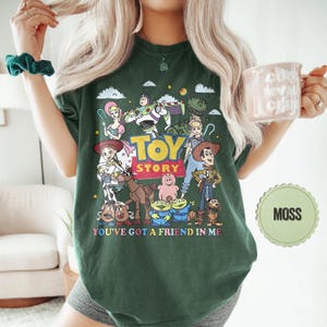 May include: A dark green t-shirt featuring a colorful graphic of characters from the animated film "Toy Story." The design includes the words "Toy Story" and "You've Got a Friend in Me."
