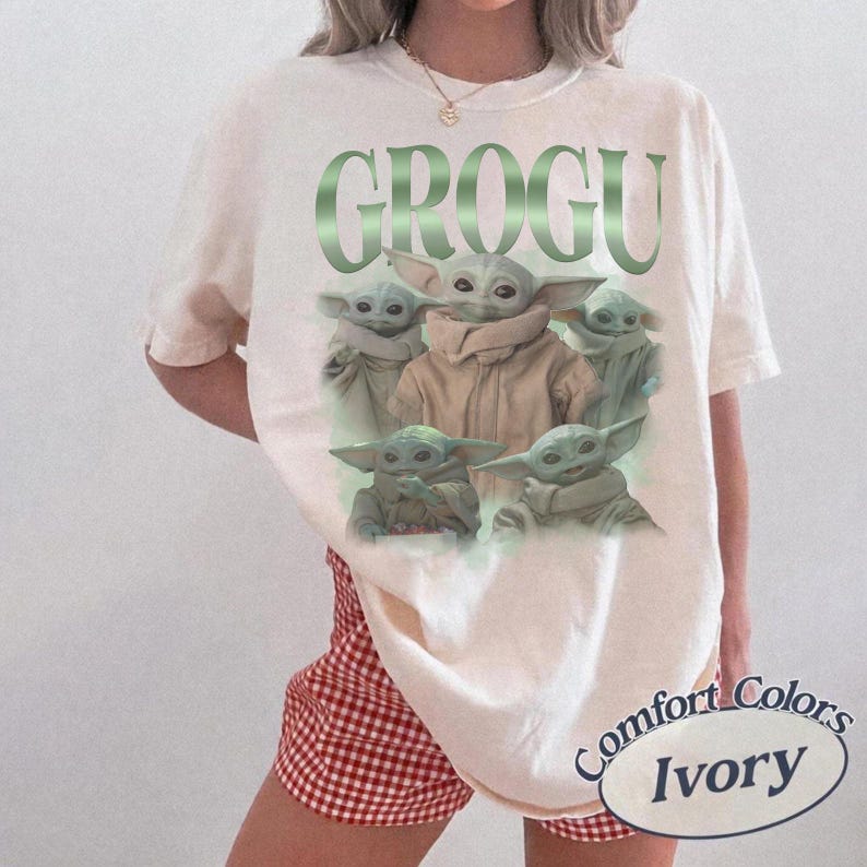 Op de afbeelding: Ivoor kleurig T-shirt met een afbeelding van Grogu, met het woord "GROGU" in groen boven de afbeelding. De afbeelding bevat meerdere afbeeldingen van Grogu. Het shirt is van Comfort Colors.