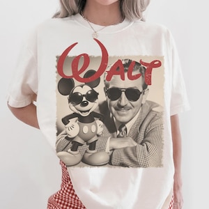 May include: White t-shirt featuring a sepia-toned image of Walt Disney and Mickey Mouse wearing sunglasses. The word "Walt" is written in red script above the image. The shirt is a casual, unisex style.
