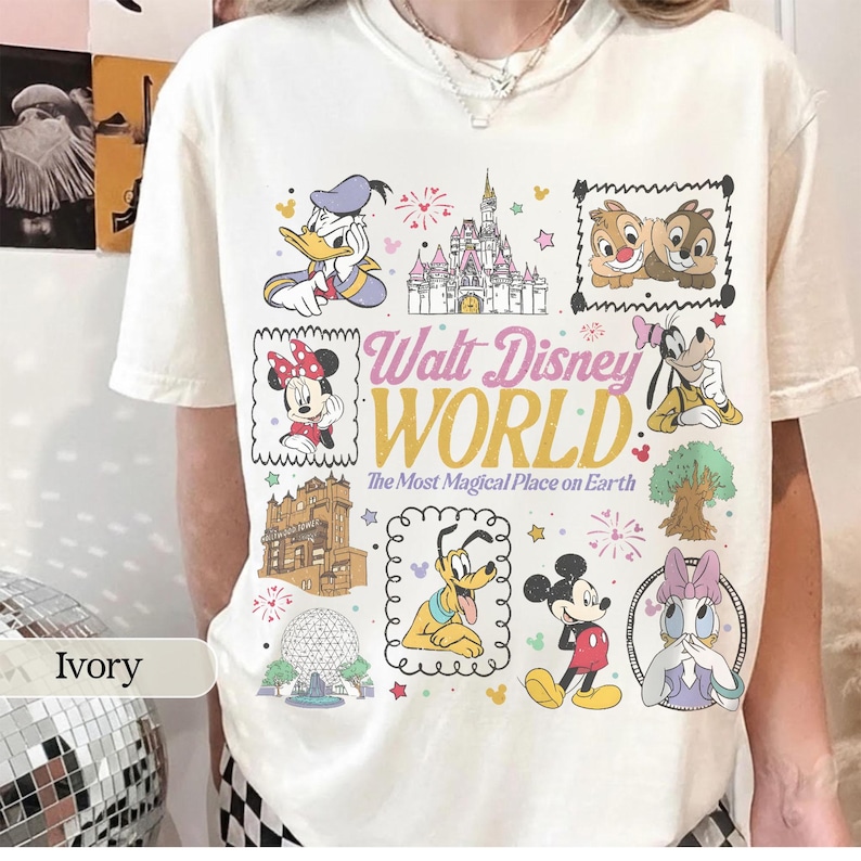 K&ouml;nnte beinhalten: Elfenbeinfarbenes T-Shirt mit einer farbenfrohen Grafik verschiedener Disney-Charaktere und Wahrzeichen, darunter Micky Maus, Minnie Maus und Cinderella's Schloss. Der Text "Walt Disney World" ist prominent dargestellt.