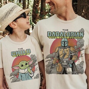K&ouml;nnte beinhalten: Zwei cremefarbene T-Shirts mit Grafikdrucken. Ein Shirt zeigt eine gr&uuml;ne Figur mit dem Text THE CHILD &uuml;ber einer roten Sonne. Das andere Shirt zeigt eine gepanzerte Figur mit dem Text THE DADALORIAN &uuml;ber einer roten Sonne.