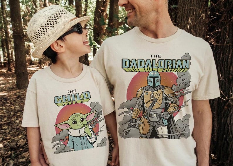 K&ouml;nnte beinhalten: Zwei cremefarbene T-Shirts mit Grafikdrucken. Ein Shirt zeigt eine gr&uuml;ne Figur mit dem Text THE CHILD &uuml;ber einer roten Sonne. Das andere Shirt zeigt eine gepanzerte Figur mit dem Text THE DADALORIAN &uuml;ber einer roten Sonne.
