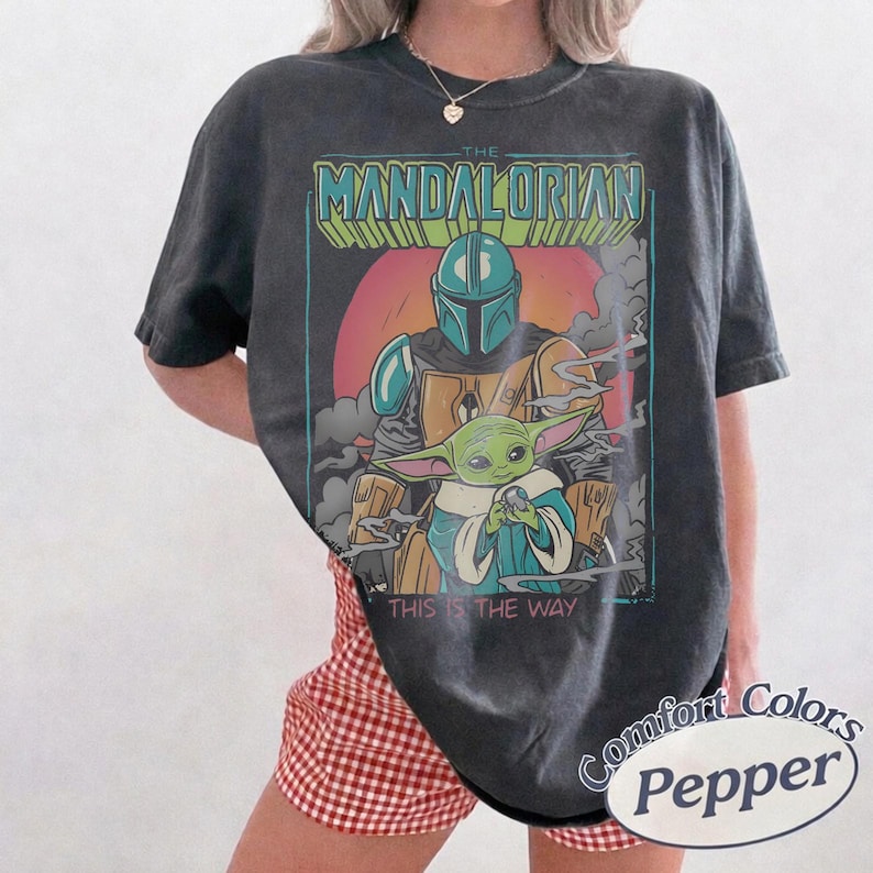 May include: A charcoal gray short-sleeve t-shirt featuring a graphic print of The Mandalorian and Grogu. The design includes the text THE MANDALORIAN and THIS IS THE WAY, set against a circular orange background with gray smoke elements.