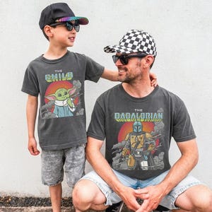 K&ouml;nnte beinhalten: Zwei anthrazitfarbene T-Shirts mit Grafikdrucken. Ein Shirt zeigt eine gr&uuml;ne Figur mit dem Text THE CHILD &uuml;ber einer roten Sonne. Das andere Shirt zeigt eine maskierte, gepanzerte Figur mit dem Text THE DADALORIAN &uuml;ber einer roten Sonne.