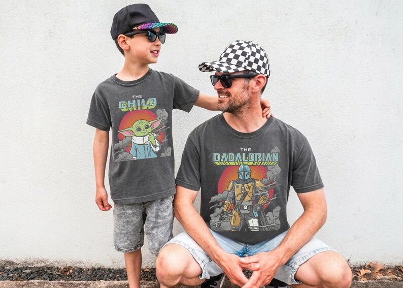 K&ouml;nnte beinhalten: Zwei anthrazitfarbene T-Shirts mit Grafikdrucken. Ein Shirt zeigt eine gr&uuml;ne Figur mit dem Text THE CHILD &uuml;ber einer roten Sonne. Das andere Shirt zeigt eine maskierte, gepanzerte Figur mit dem Text THE DADALORIAN &uuml;ber einer roten Sonne.