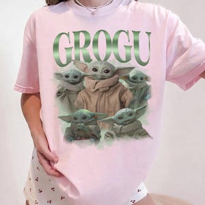 Op de afbeelding: Lichtroze T-shirt met het woord "GROGU" in groen en een afbeelding van Grogu uit The Mandalorian. De afbeelding toont Grogu in verschillende poses. Het shirt heeft korte mouwen.