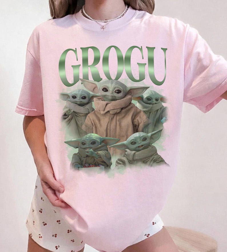 Op de afbeelding: Lichtroze T-shirt met het woord "GROGU" in groen en een afbeelding van Grogu uit The Mandalorian. De afbeelding toont Grogu in verschillende poses. Het shirt heeft korte mouwen.
