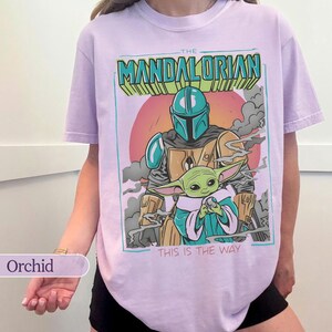 May include: A light purple t-shirt featuring a graphic print of The Mandalorian and Grogu. The design includes the text THE MANDALORIAN at the top and THIS IS THE WAY at the bottom, set against a circular orange sun and gray smoke clouds.