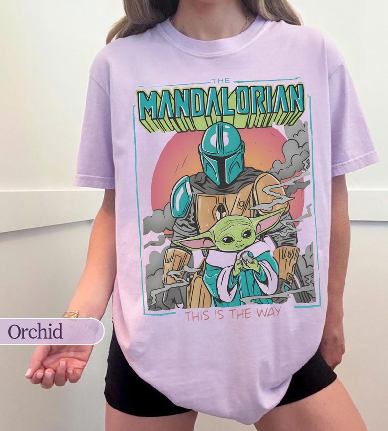 May include: A light purple t-shirt featuring a graphic print of The Mandalorian and Grogu. The design includes the text THE MANDALORIAN at the top and THIS IS THE WAY at the bottom, set against a circular orange sun and gray smoke clouds.