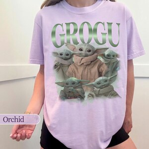 Op de afbeelding: Een lichtpaars t-shirt met het woord "GROGU" in groene metallic letters. Het shirt toont een afbeelding van Grogu, ook bekend als Baby Yoda, uit de Star Wars-serie. Het t-shirt is lichtpaars van kleur.